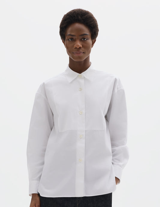 Cropped body image, front view of model wearing white oversized cotton shirt with pointed stand collar, mother of pearl buttons on placket, double-layer bib front and curved hem. Styled with black jeans.