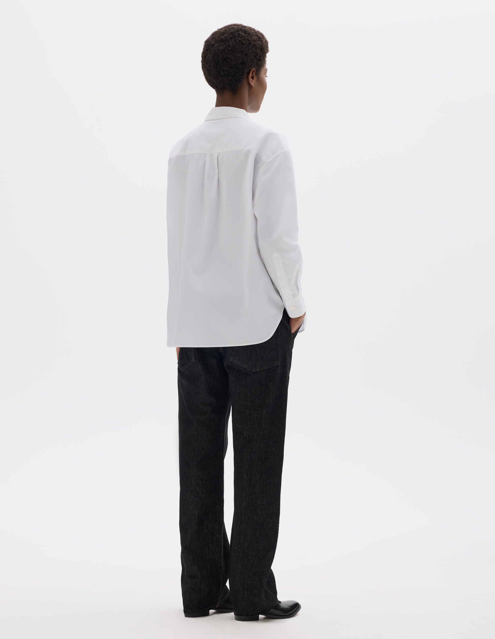 Full body image, back view of model wearing white oversized cotton shirt with narrow shoulder yoke and back pleat, mother of pearl buttons on cuffs and curved hem. Styled with black jeans and black leather shoes.