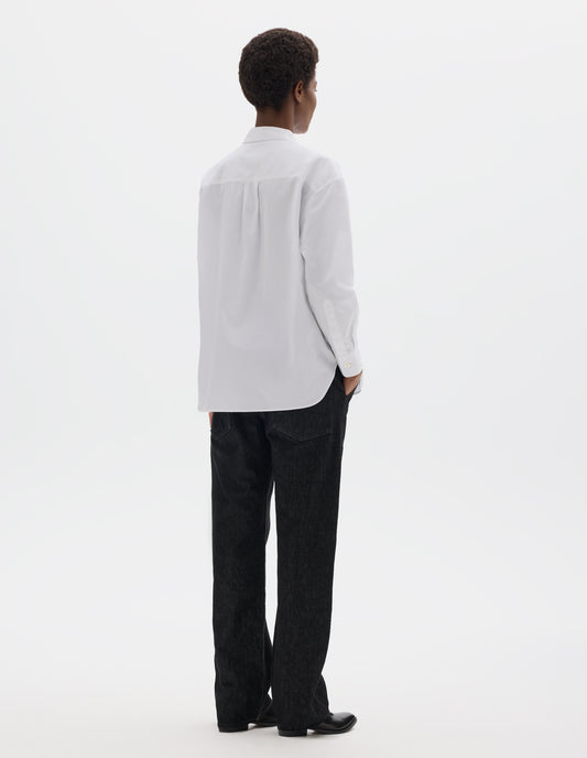Full body image, back view of model wearing white oversized cotton shirt with narrow shoulder yoke and back pleat, mother of pearl buttons on cuffs and curved hem. Styled with black jeans and black leather shoes.