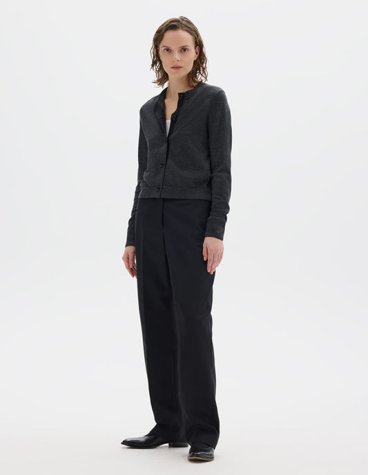 Full body image, front view of model wearing dark grey merino cardigan with fully fashioned sleeves, horn buttons on concealed placket finished with grosgrain, and ribbed neck, cuffs and hem. Styled over white top and with black trousers and black leather shoes.