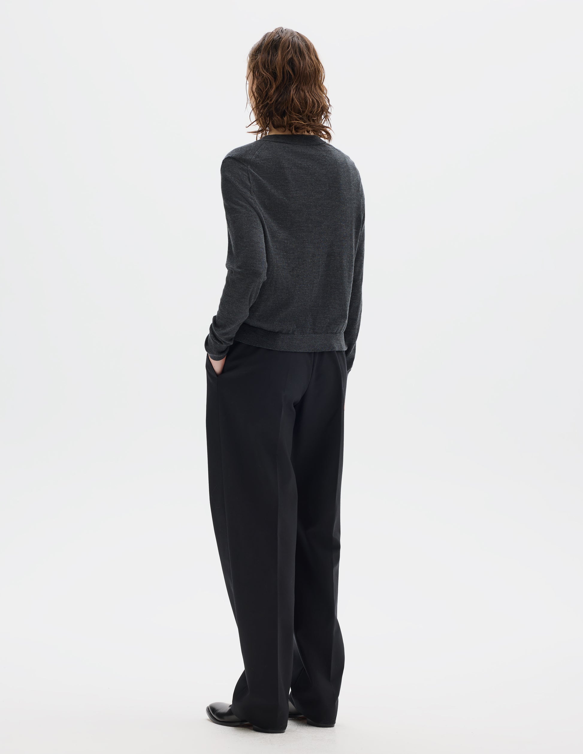 Full body image, back view of model wearing dark grey merino cardigan with fully fashioned sleeves, French shoulder offset on back seam, and ribbed neck, cuffs and hem. Styled with black trousers and black leather shoes.