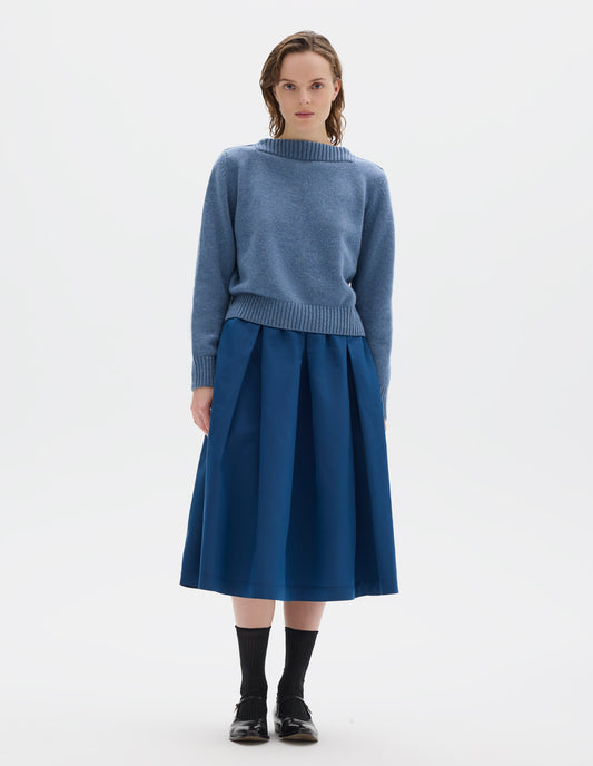 Full body image, front view of model wearing cropped light blue merino cashmere fully fashioned jumper with higher square neckline and with wide rib detailing on neck, cuffs and hem. Styled with blue skirt, black socks and black leather shoes.