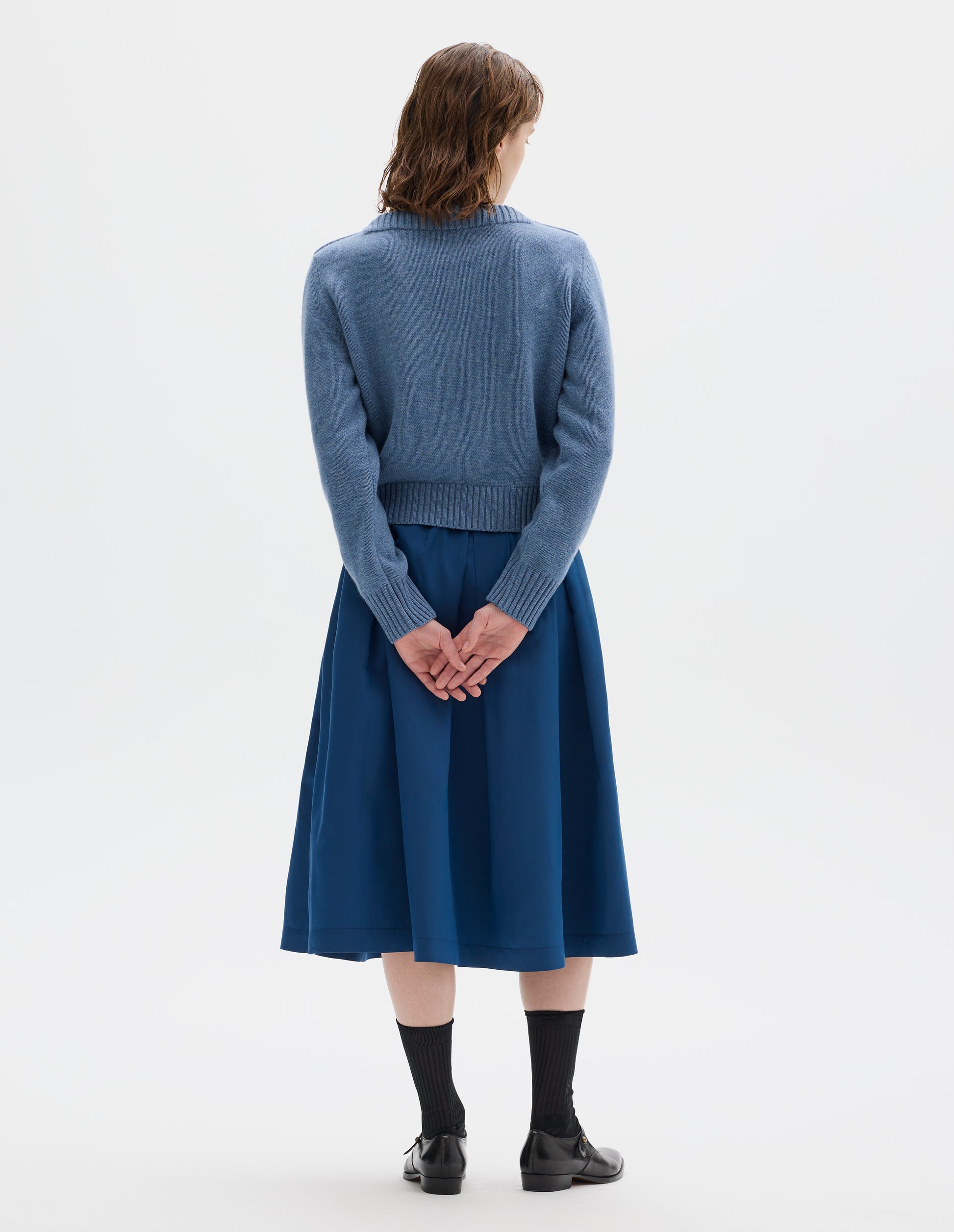 Full body image, back view of model wearing cropped light blue merino cashmere fully fashioned jumper with higher square neckline and with wide rib detailing on neck, cuffs and hem. Styled with blue skirt, black socks and black leather shoes.