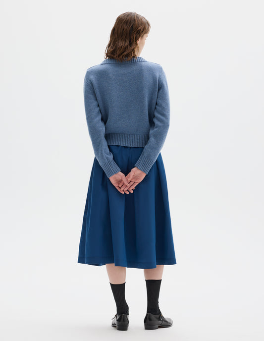 Full body image, back view of model wearing cropped light blue merino cashmere fully fashioned jumper with higher square neckline and with wide rib detailing on neck, cuffs and hem. Styled with blue skirt, black socks and black leather shoes.