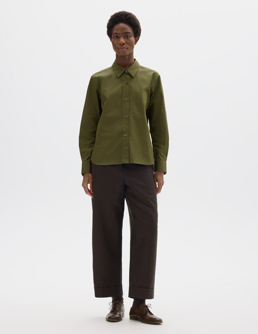 Full body image, front view of model wearing green cotton shirt with mother of pearl buttons on raised placket, chest patch pocket and curved hem. Styled with brown trousers and brown leather brogues.