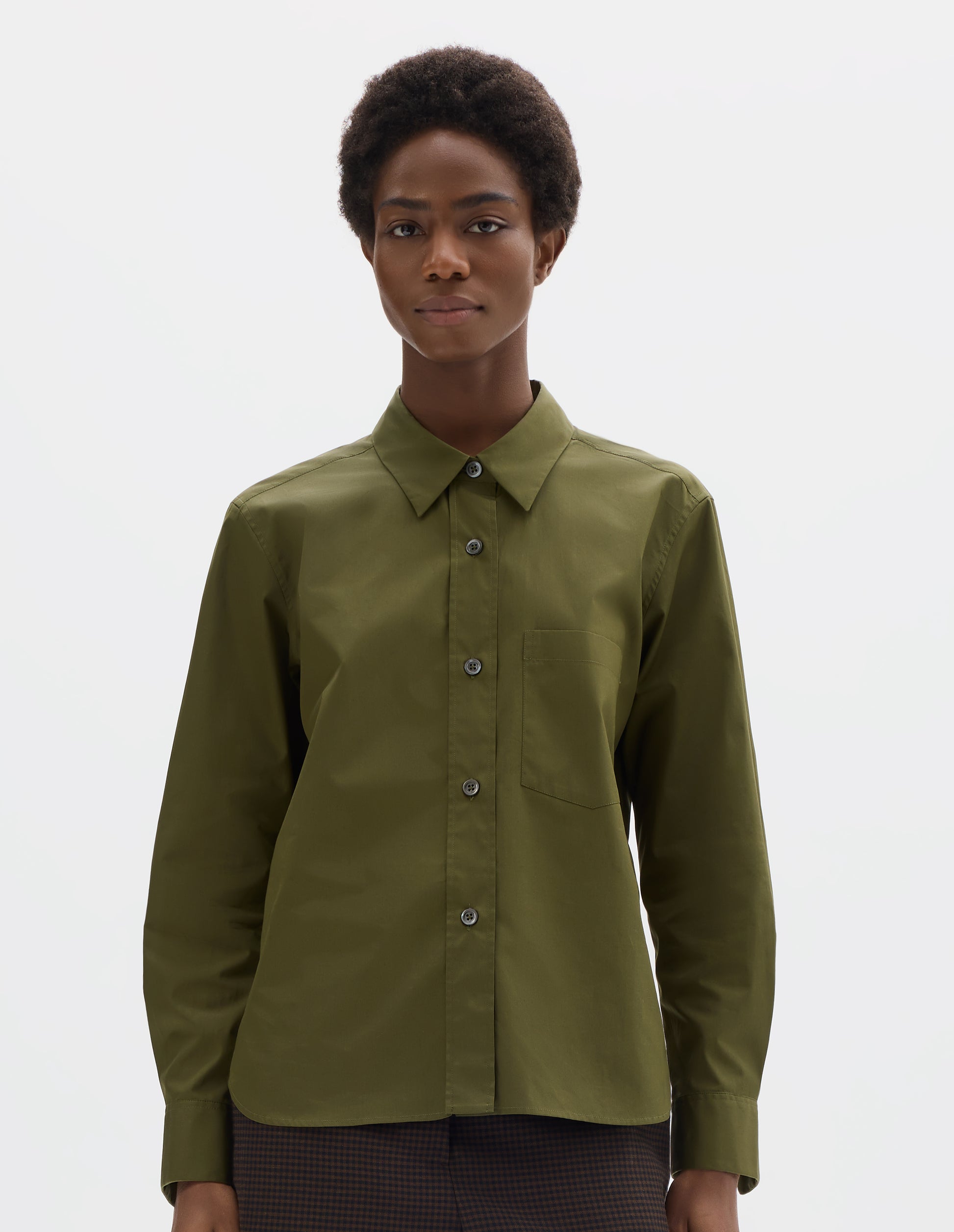 Cropped body image, front view of model wearing green cotton shirt with mother of pearl buttons on raised placket, chest patch pocket and curved hem. Styled with brown trousers.