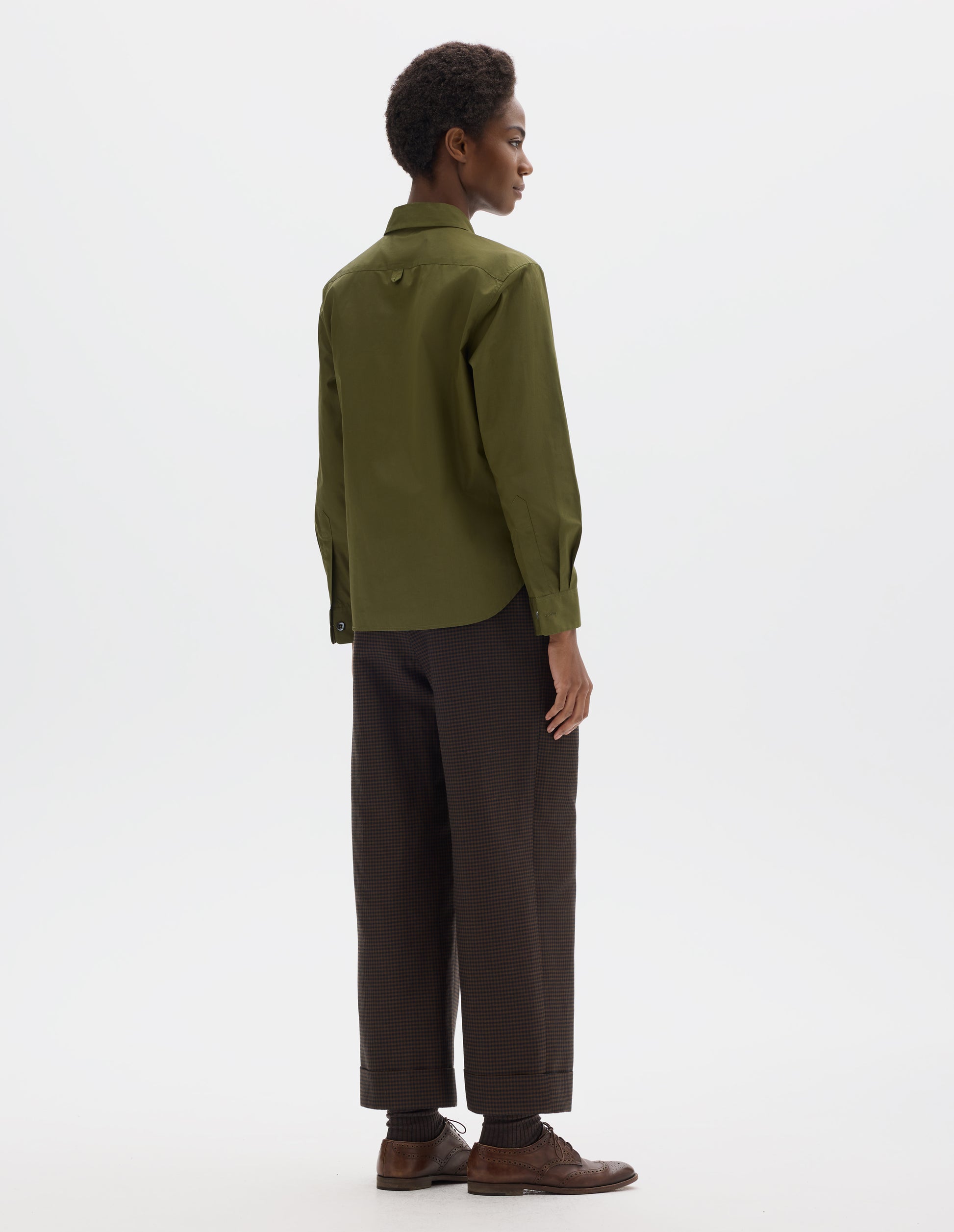 Full body image, back view of model wearing green cotton shirt with hanger loop detail on back yoke, mother of pearl buttons on cuffs and curved hem.  Styled with brown trousers and brown leather brogues.