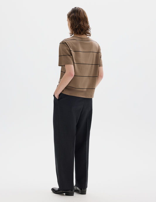 Full body image, back view of model wearing relaxed beige jersey t-shirt with black stripes, boxy cut and with crew neck. Styled with dark grey trousers and black leather shoes.