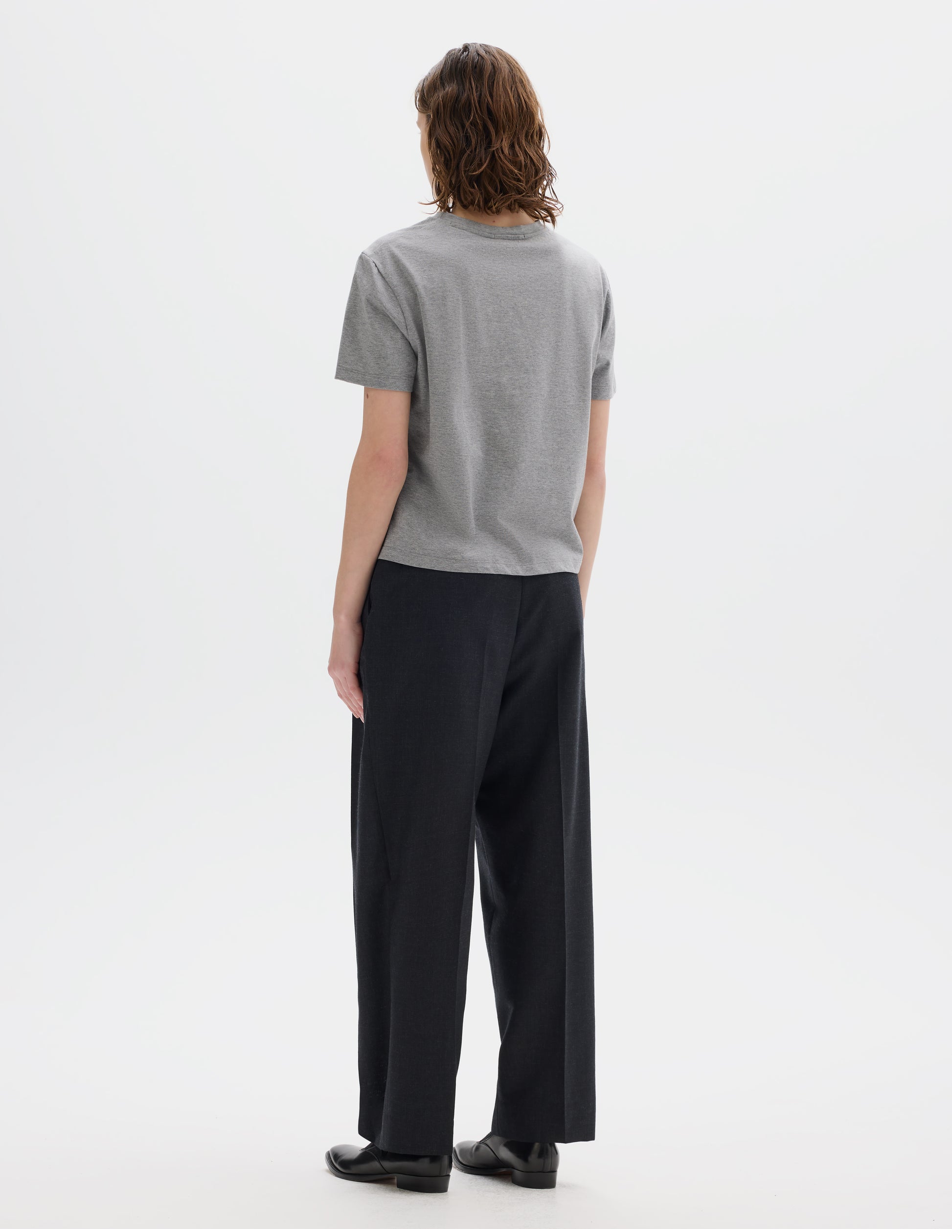 Full body image, back view of model wearing grey relaxed cotton t-shirt with crew neck and boxy cut. Styled with dark grey trousers and black leather shoes.