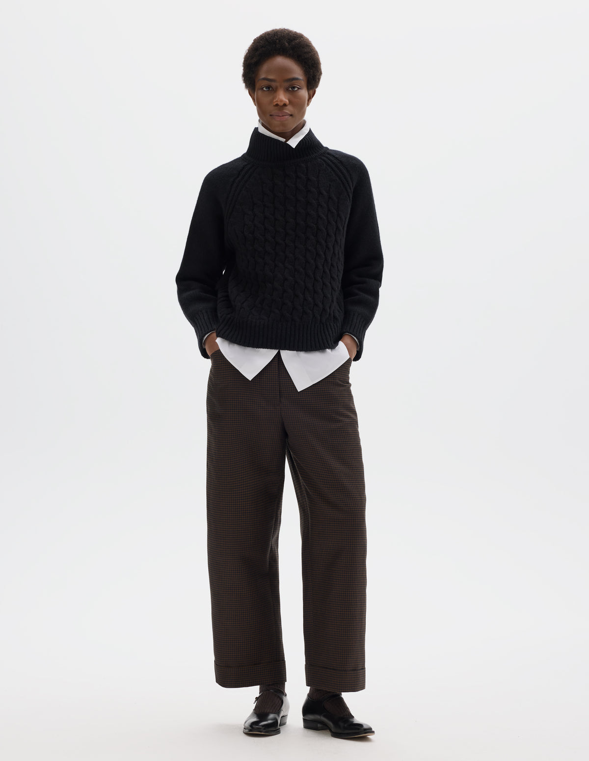 Full body image, front view of model wearing relaxed black merino jumper with ribbed high neck collar, front cable design, raglan sleeves with ribbed side seams, cuffs and hem. Styled over white shirt, and with brown check trousers and black leather shoes.