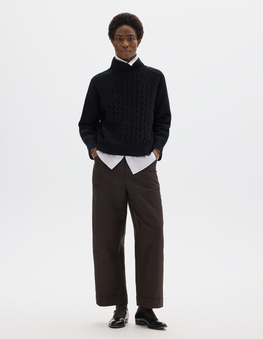 Full body image, front view of model wearing relaxed black merino jumper with ribbed high neck collar, front cable design, raglan sleeves with ribbed side seams, cuffs and hem. Styled over white shirt, and with brown check trousers and black leather shoes.
