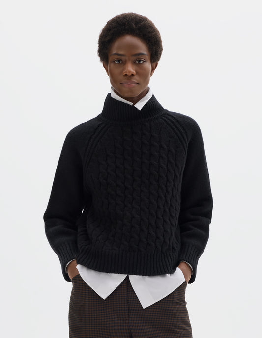 Cropped body image, front view of model wearing relaxed black merino jumper with ribbed high neck collar, front cable design, raglan sleeves with ribbed side seams, cuffs and hem. Styled over white shirt, and with brown check trousers.
