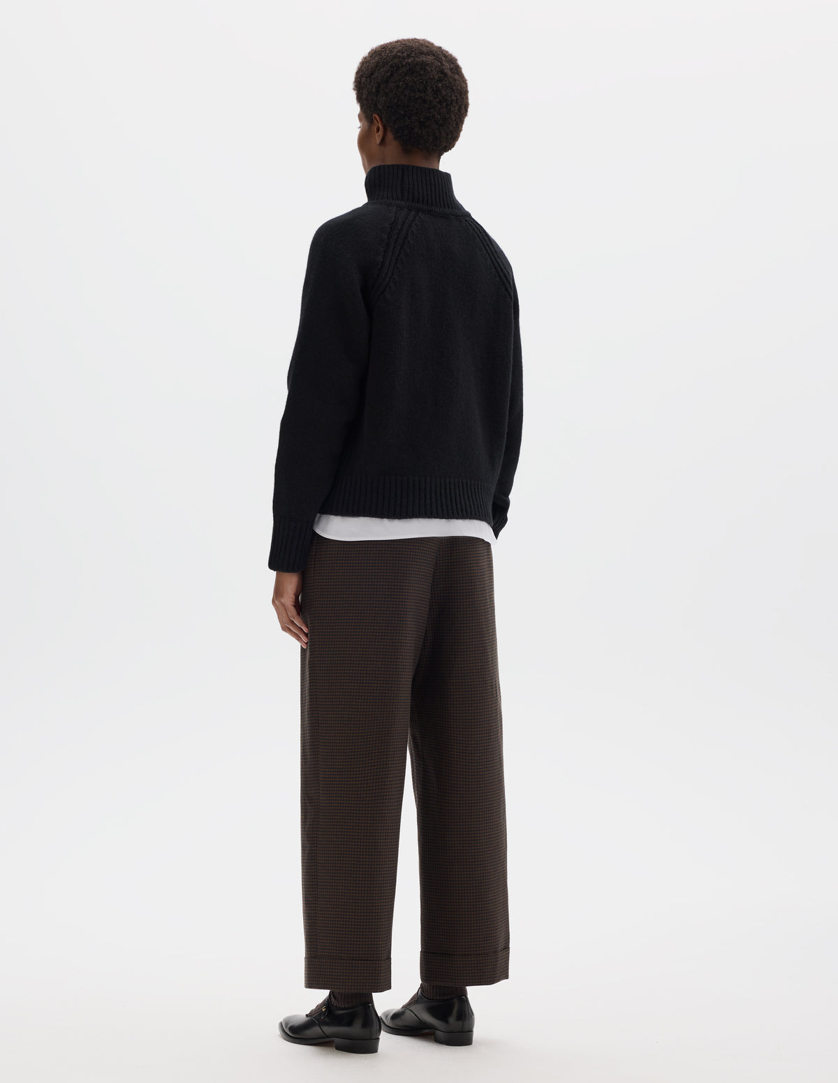Full body image, back view of model wearing relaxed black merino jumper with plain knitted back, raglan sleeves with ribbed side seams and ribbed high neck collar, cuffs and hem. Styled over white shirt, and with brown check trousers and black leather shoes.
