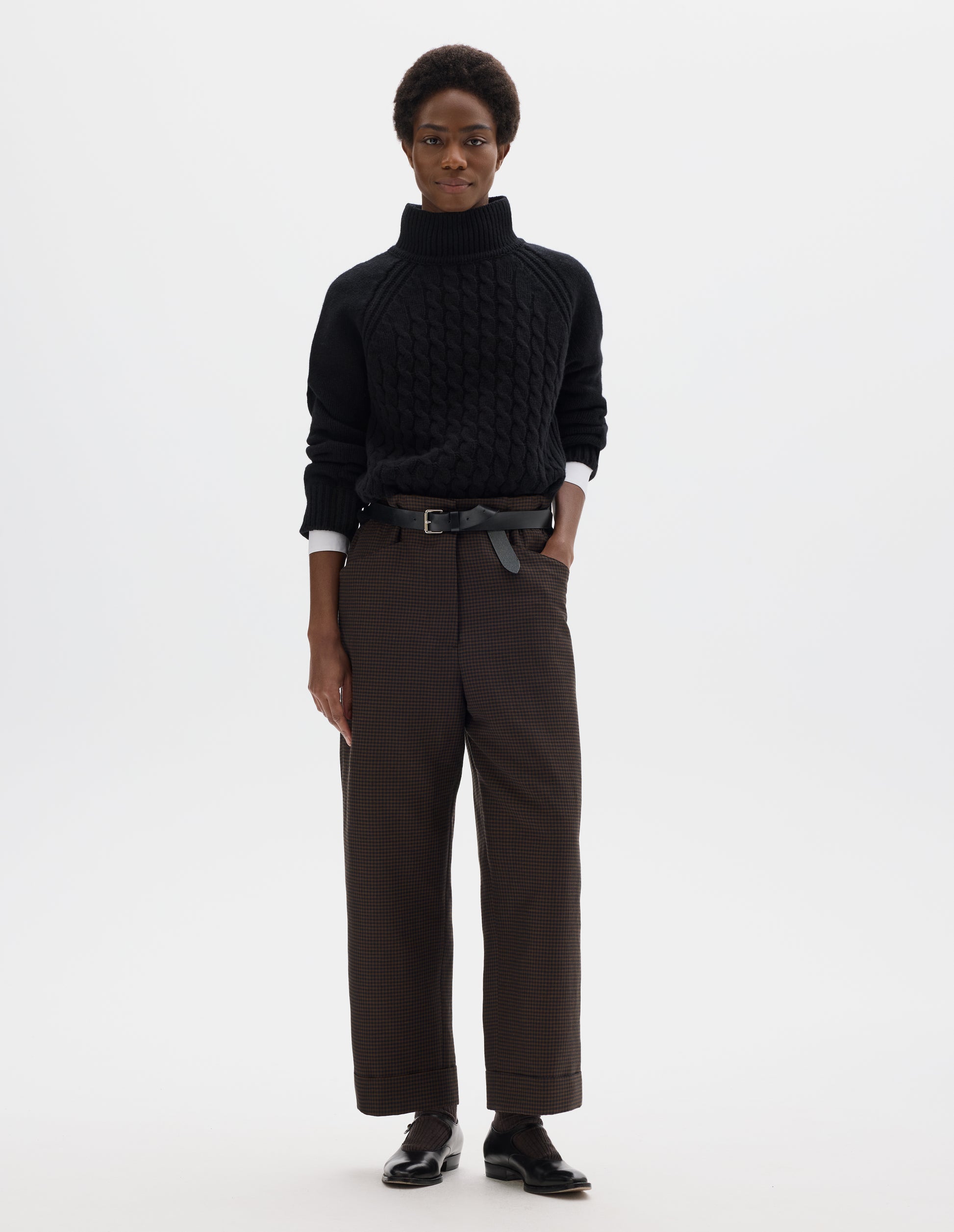 Full body image, front view of model wearing cropped length relaxed dark blue and dark brown linen wool gingham trousers with zip fly and horn button fastening on elasticated waistband, frog pockets and stitch-down turn-up hem. Styled with white shirt, black jumper, black leather belt and black leather shoes.