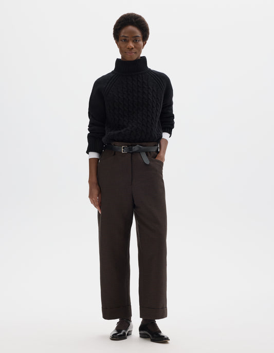 Full body image, front view of model wearing cropped length relaxed dark blue and dark brown linen wool gingham trousers with zip fly and horn button fastening on elasticated waistband, frog pockets and stitch-down turn-up hem. Styled with white shirt, black jumper, black leather belt and black leather shoes.