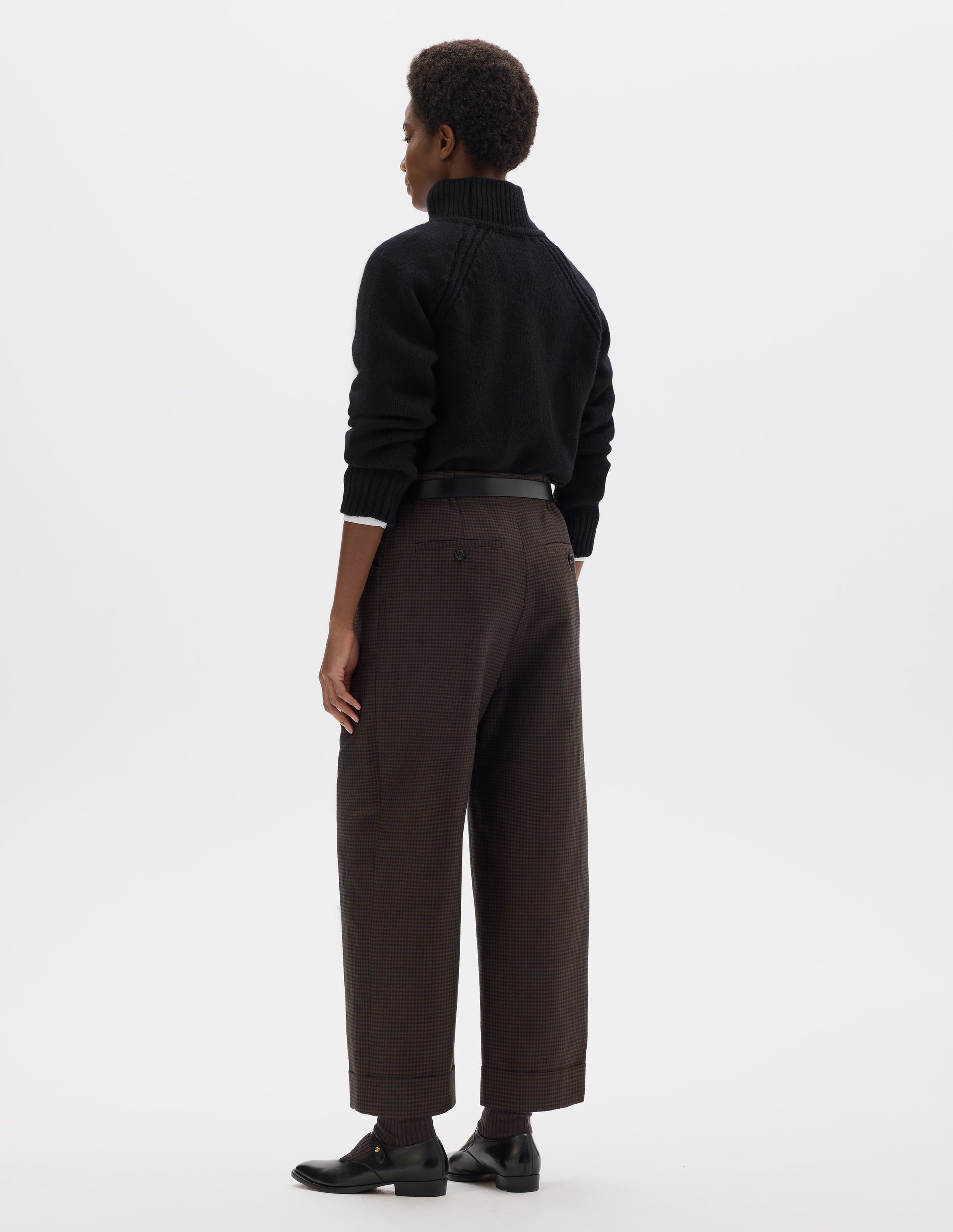 Full body image, back view of model wearing cropped length relaxed dark blue and dark brown linen wool gingham trousers with elasticated waistband, horn button-through jet back pockets and stitch-down turn-up hem. Styled with white shirt, black jumper, black leather belt and black leather shoes.