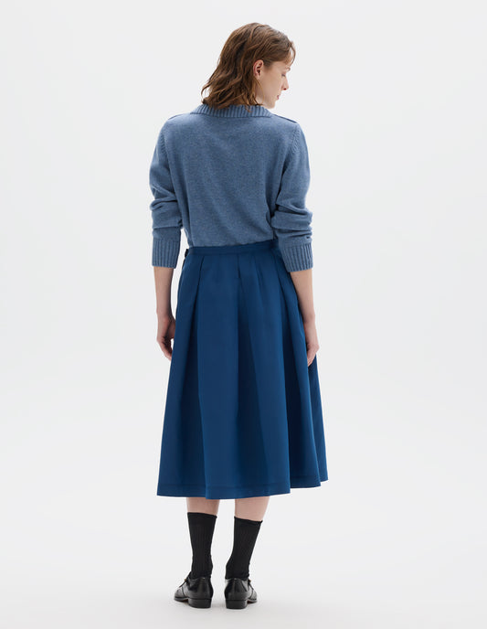 Full body image, back view of model wearing midi length blue cotton silk skirt with narrow waistband, concealed side zip with hook and bar fastening, box pleats with darts and concealed side seam pockets. Styled with light blue jumper, black socks and black leather shoes.