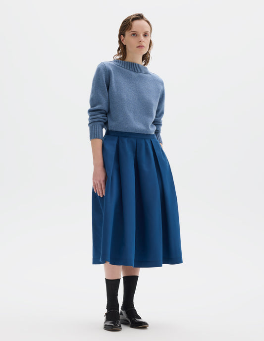 Full body image, front view of model wearing midi length blue cotton silk skirt with narrow waistband, concealed side zip with hook and bar fastening, box pleats with darts and concealed side seam pockets. Styled with light blue jumper, black socks and black leather shoes.
