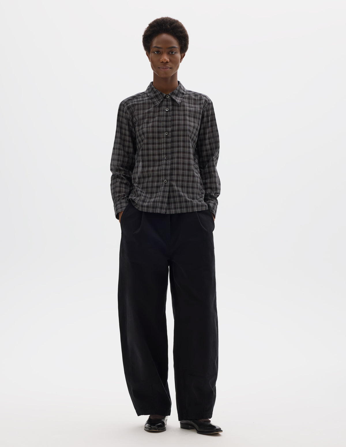 Full body image, front view of model wearing black and white cotton check shirt with mother of pearl buttons on placket, stepped hem with curved front and straight-cut back. Styled with black trousers and black leather shoes.