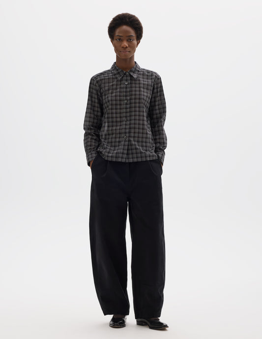 Full body image, front view of model wearing black and white cotton check shirt with mother of pearl buttons on placket, stepped hem with curved front and straight-cut back. Styled with black trousers and black leather shoes.