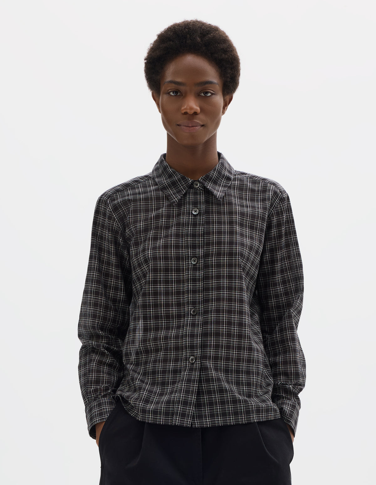 Cropped body image, front view of model wearing black and white cotton check shirt with mother of pearl buttons on placket, stepped hem with curved front and straight-cut back. Styled with black trousers.