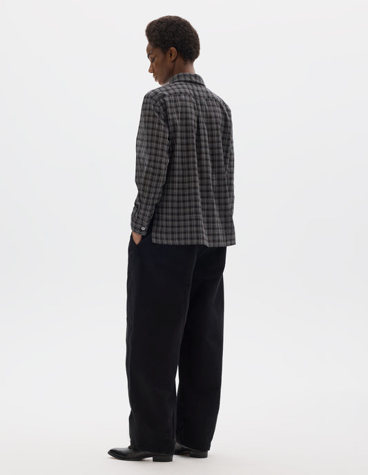 Full body image, back view of model wearing black and white cotton check shirt with pleat on back yoke, mother of pearl buttons cuffs and stepped hem. Styled with black trousers and black leather shoes.