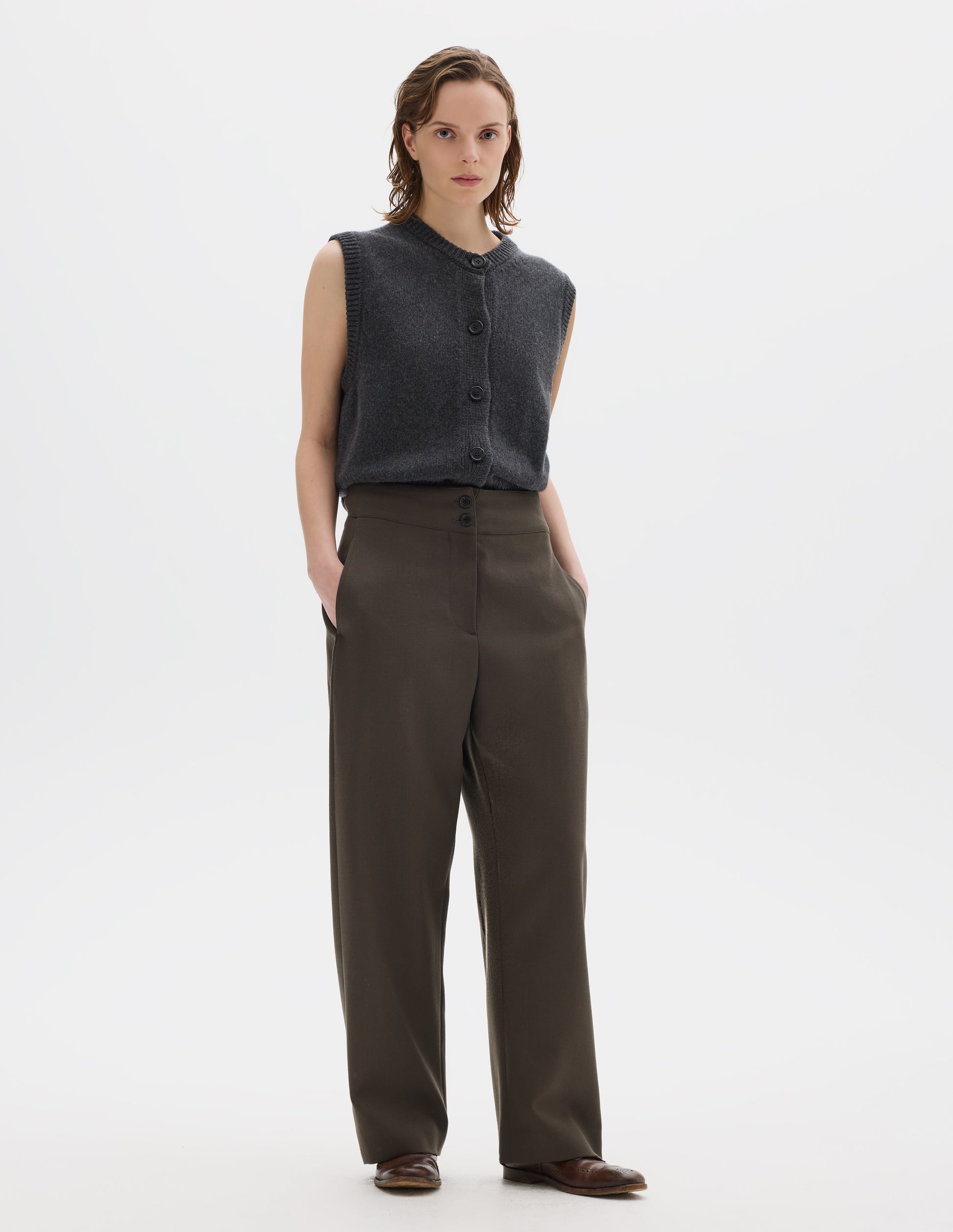 Full body image, front view of model wearing high waisted brown wool trousers with double horn button fastening and concealed side pockets with stitch detailing. Styled with grey knitted gilet and brown leather brogues.