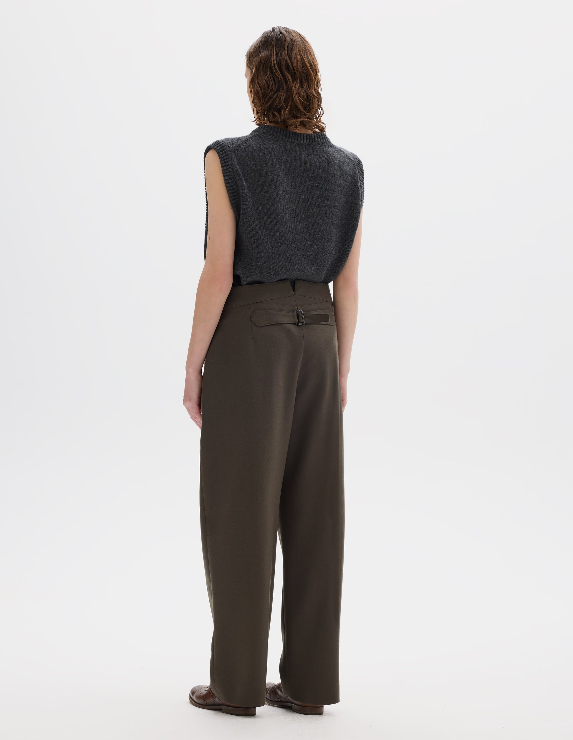 Full body image, back view of model wearing high waisted brown wool trousers with back cinch. Styled with grey knitted gilet and brown leather brogues.
