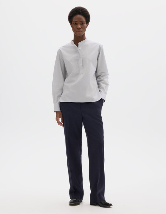 Full body image, front view of model wearing relaxed white collarless cotton shirt with dark blue stripes, mother of pearl buttons on placket with box stitch detailing and curved stepped hem. Styled with dark blue trousers and black leather shoes.