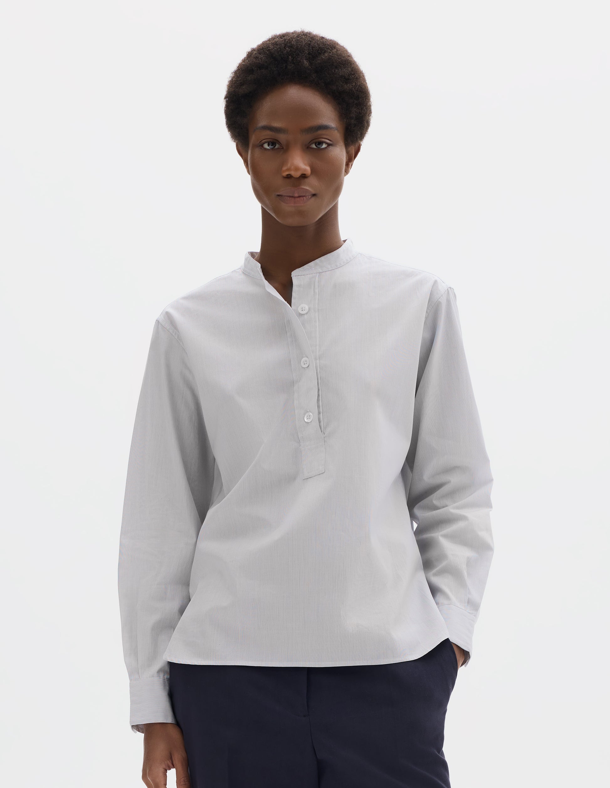 Cropped body image, front view of model wearing relaxed white collarless cotton shirt with dark blue stripes, mother of pearl buttons on placket with box stitch detailing and curved stepped hem. Styled with dark blue trousers.