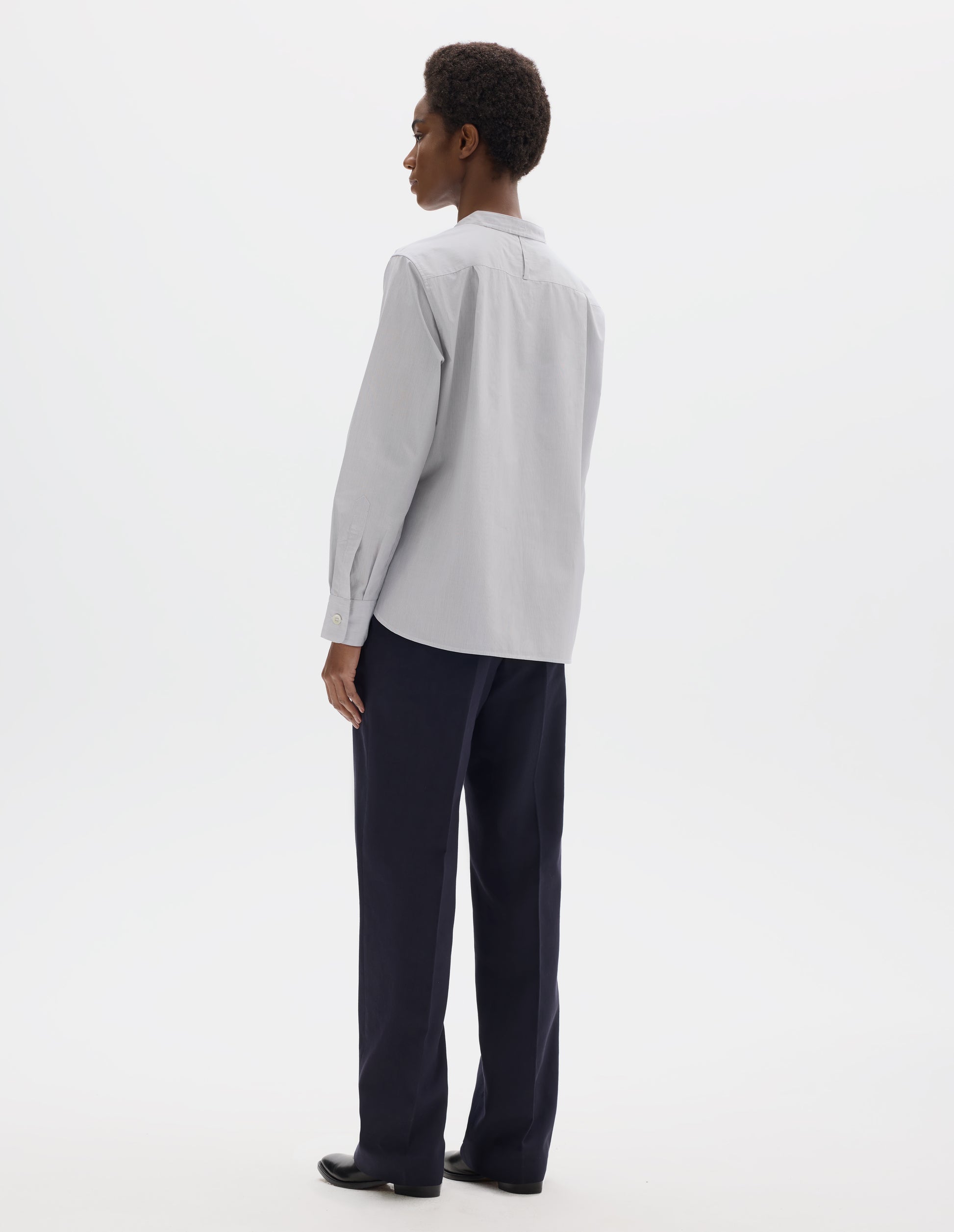 Full body image, back view of model wearing relaxed white collarless cotton shirt with dark blue stripes, narrow shoulder yoke with hanger loop, curved stepped hem and mother of pearl buttons on cuffs. Styled with dark blue trousers and black leather shoes.