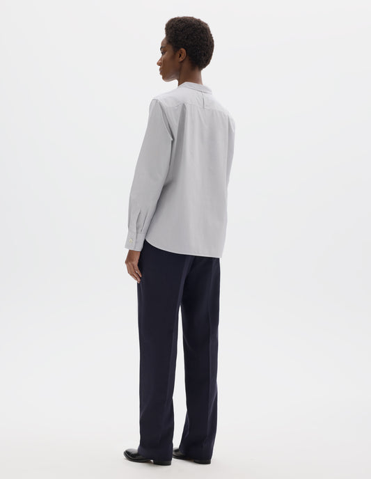 Full body image, back view of model wearing relaxed white collarless cotton shirt with dark blue stripes, narrow shoulder yoke with hanger loop, curved stepped hem and mother of pearl buttons on cuffs. Styled with dark blue trousers and black leather shoes.