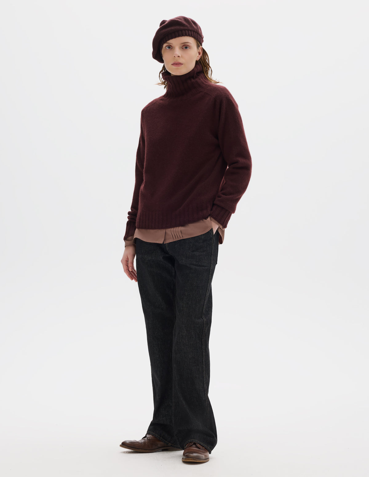Full body image, front view of model wearing seamless dark red cashmere roll neck jumper with contrast ribbing on neck, cuffs and hem. Styled over light brown shirt, and with dark brown beret, black jeans and brown leather brogues.