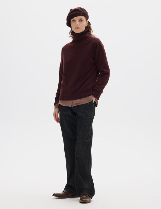 Full body image, front view of model wearing seamless dark red cashmere roll neck jumper with contrast ribbing on neck, cuffs and hem. Styled over light brown shirt, and with dark brown beret, black jeans and brown leather brogues.