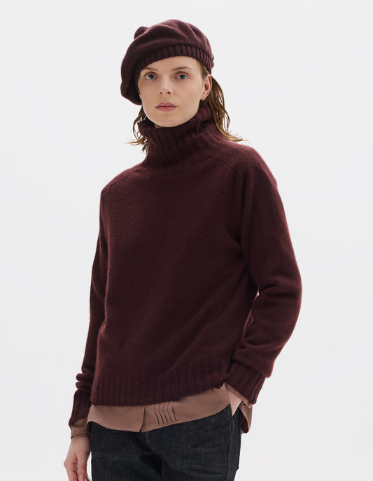 Cropped body image, front view of model wearing seamless dark red cashmere roll neck jumper with contrast ribbing on neck, cuffs and hem. Styled over light brown shirt, and with dark brown beret and black jeans.