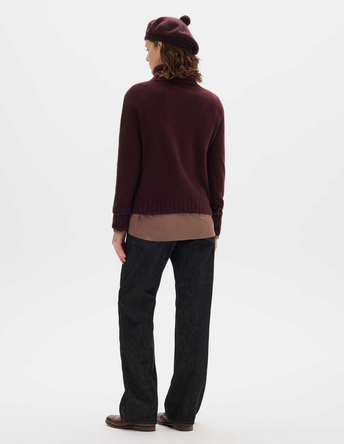 Full body image, back view of model wearing seamless dark red cashmere roll neck jumper with contrast ribbing on neck, cuffs and hem. Styled over light brown shirt, and with dark brown beret, black jeans and brown leather brogues.