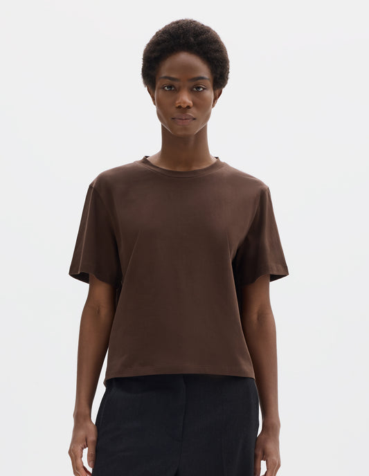 Cropped body image, front view of model wearing relaxed dark brown jersey t-shirt with crew neck and boxy cut. Styled with dark blue trousers.