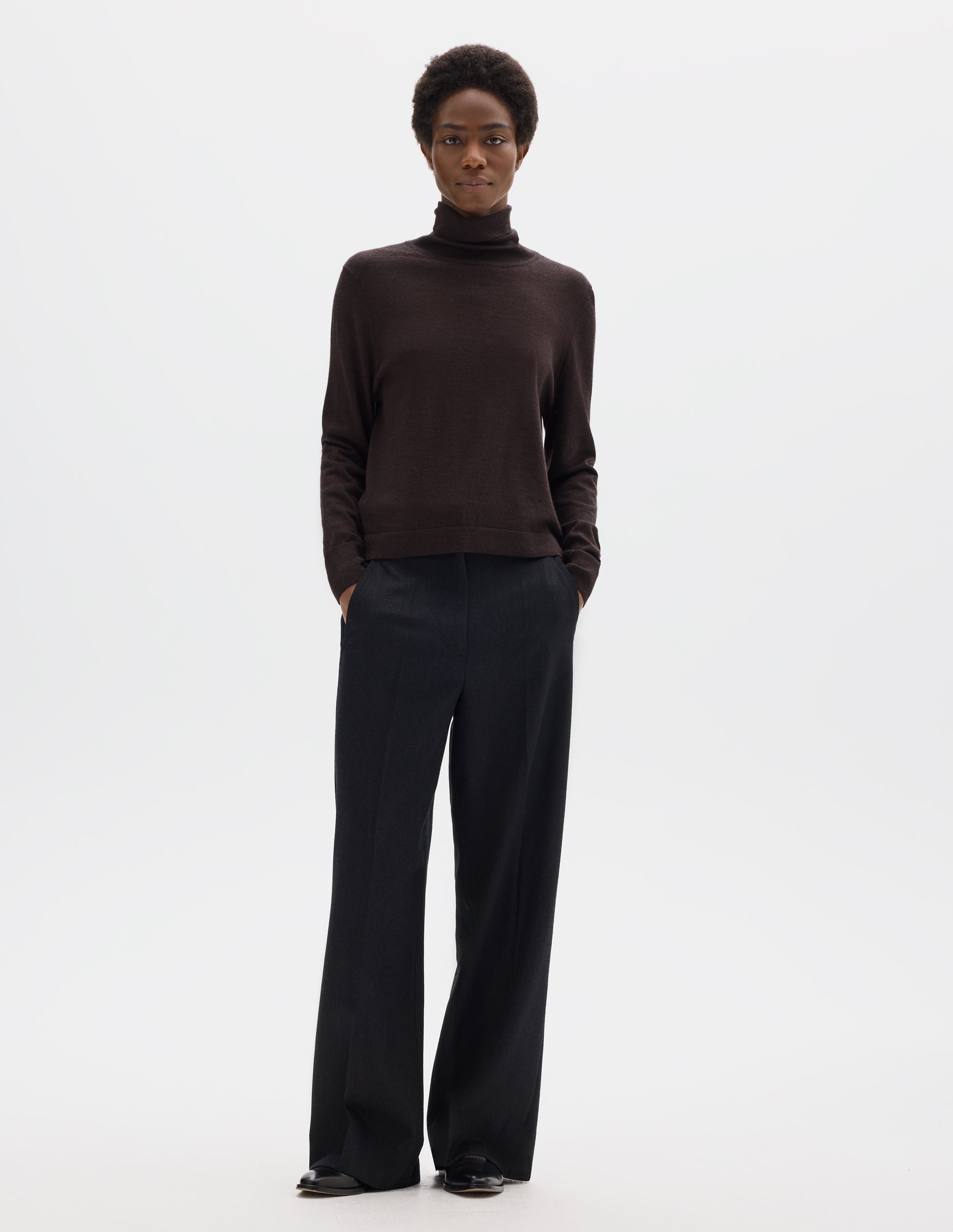 Full body image, front view of model wearing dark brown merino knitted top with fully fashioned roll neck and armhole, and with ribbed neck, sleeves, cuff and hem. Styled with black trousers and black leather shoes.