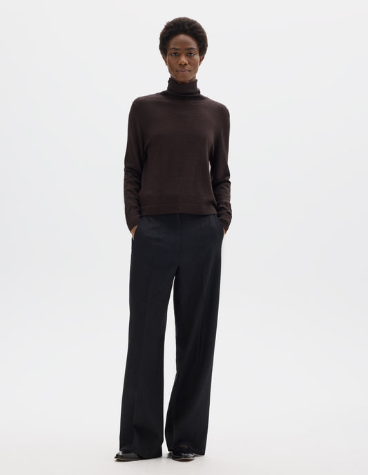 Full body image, front view of model wearing dark brown merino knitted top with fully fashioned roll neck and armhole, and with ribbed neck, sleeves, cuff and hem. Styled with black trousers and black leather shoes.