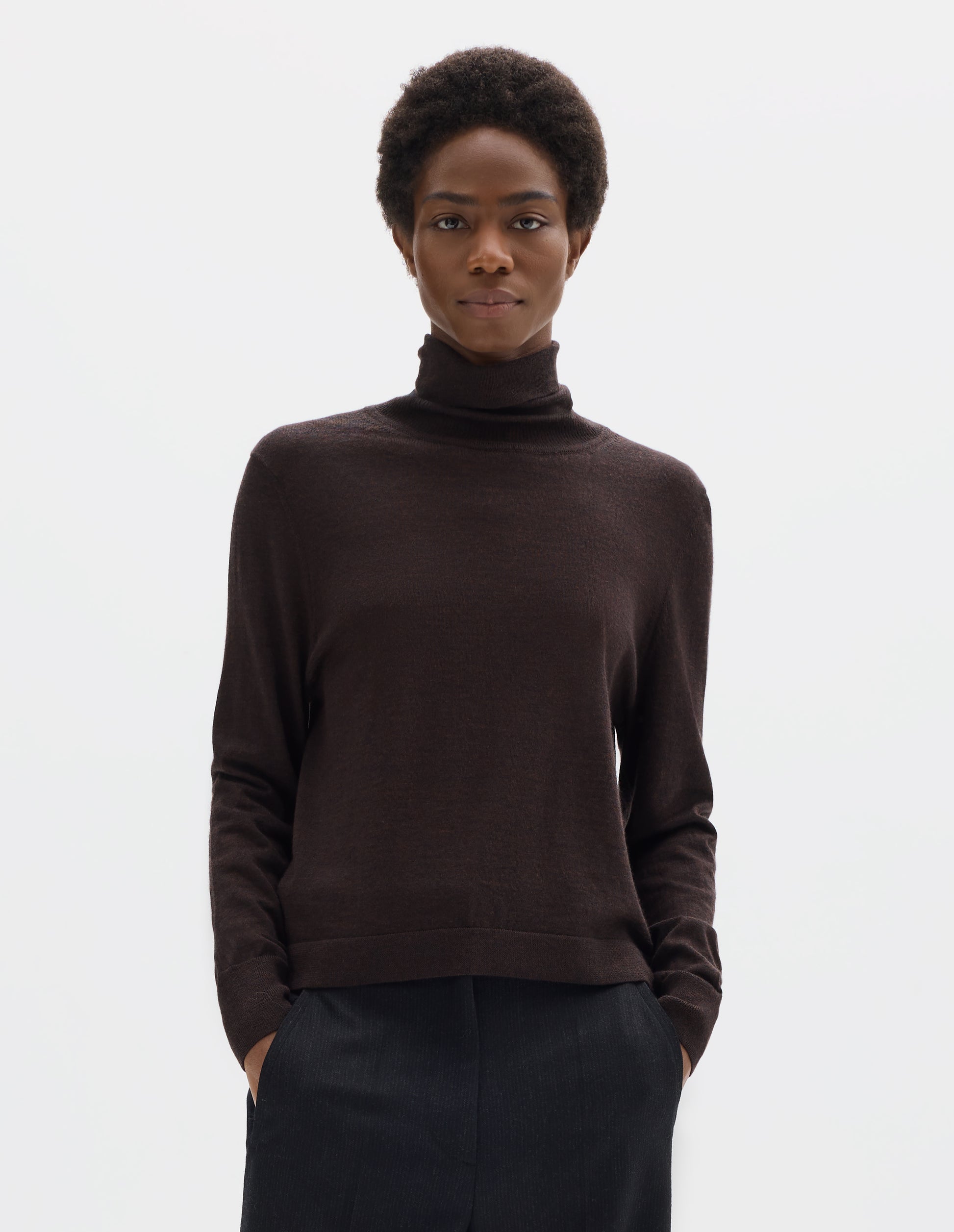 Cropped body image, front view of model wearing dark brown merino knitted top with fully fashioned roll neck and armhole, and with ribbed neck, sleeves, cuff and hem. Styled with black trousers.