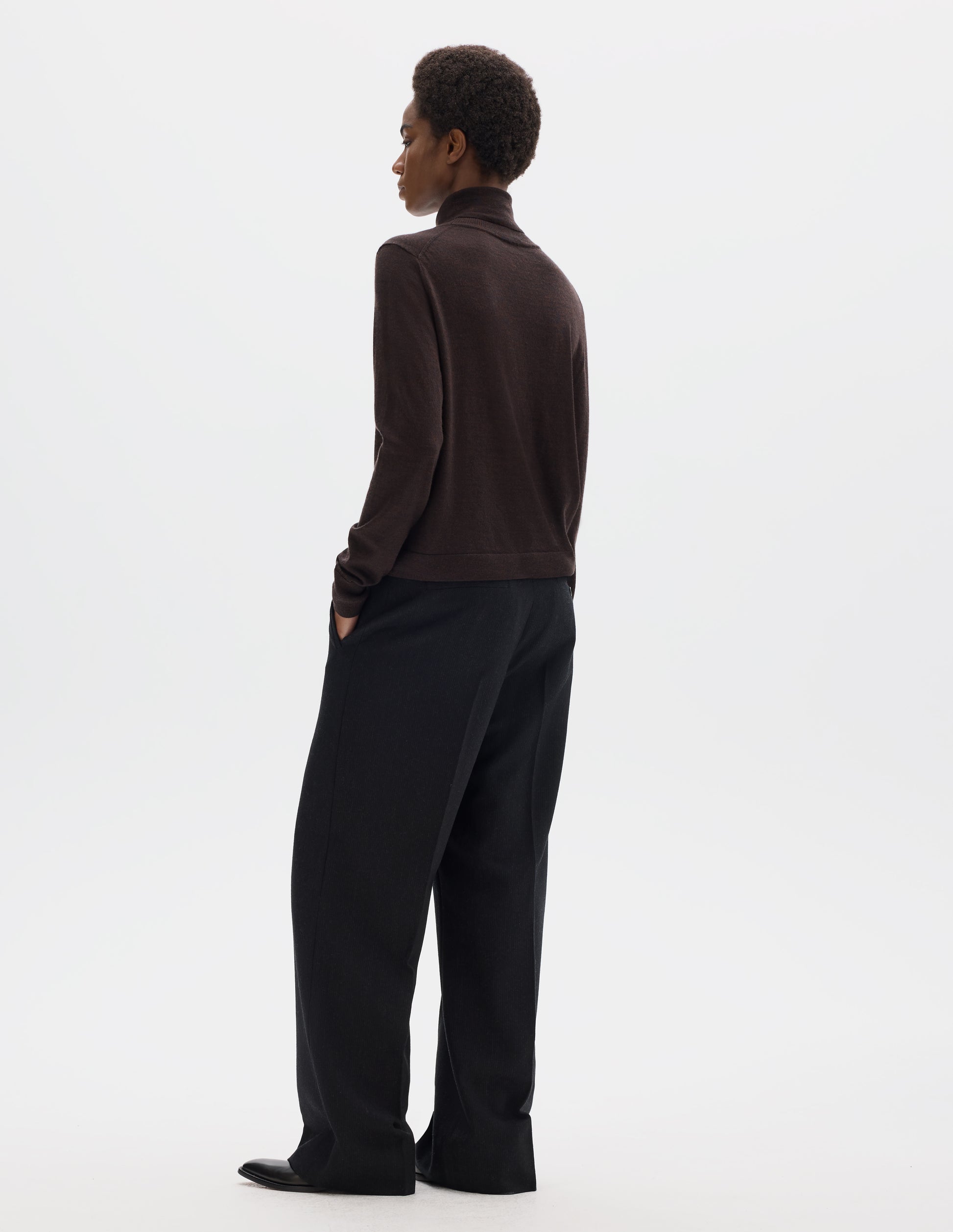 Full body image, back view of model wearing dark brown merino knitted top with fully fashioned roll neck and armhole, and with ribbed neck, sleeves, cuff and hem. Styled with black trousers and black leather shoes.