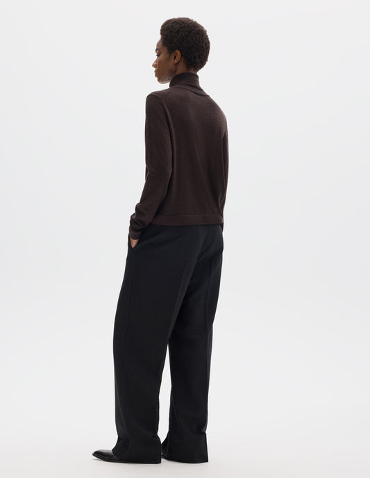 Full body image, back view of model wearing dark brown merino knitted top with fully fashioned roll neck and armhole, and with ribbed neck, sleeves, cuff and hem. Styled with black trousers and black leather shoes.