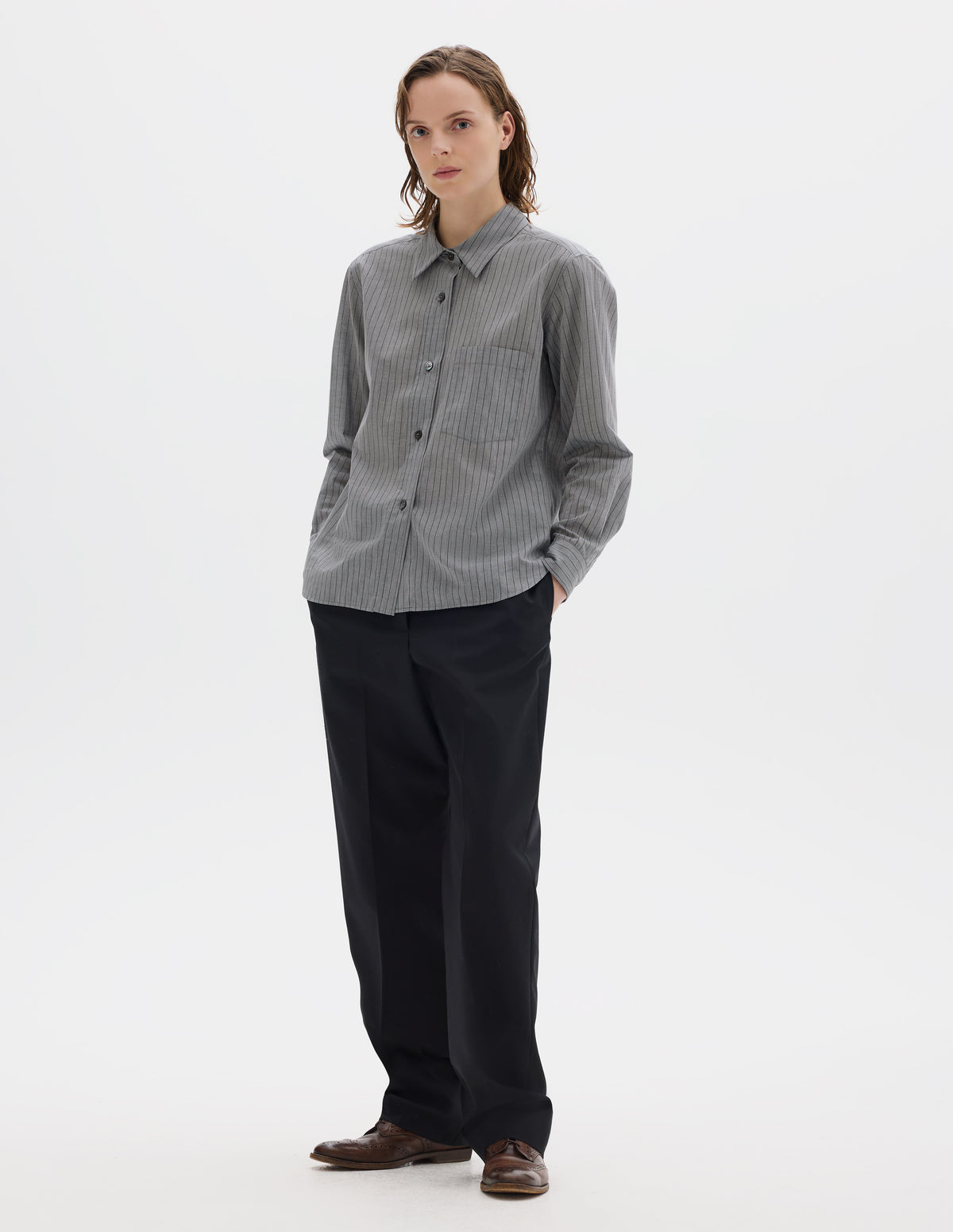 Full body image, front view of model wearing grey cotton shirt with black stipes, mother of pearl buttons on raised placket, chest patch pocket and curved hem. Styled with dark blue trousers and brown leather brogues.