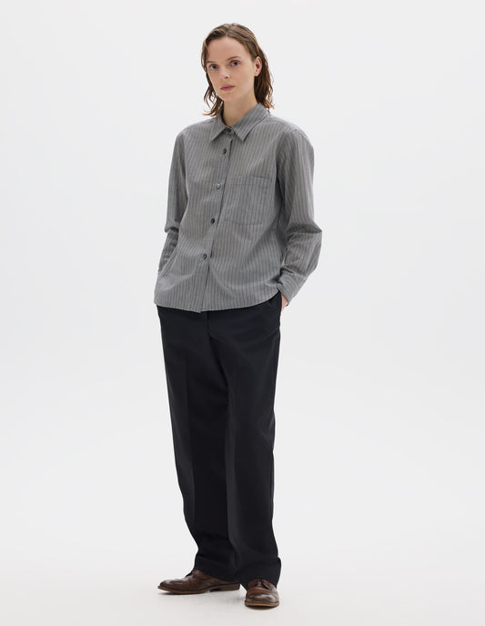 Full body image, front view of model wearing grey cotton shirt with black stipes, mother of pearl buttons on raised placket, chest patch pocket and curved hem. Styled with dark blue trousers and brown leather brogues.