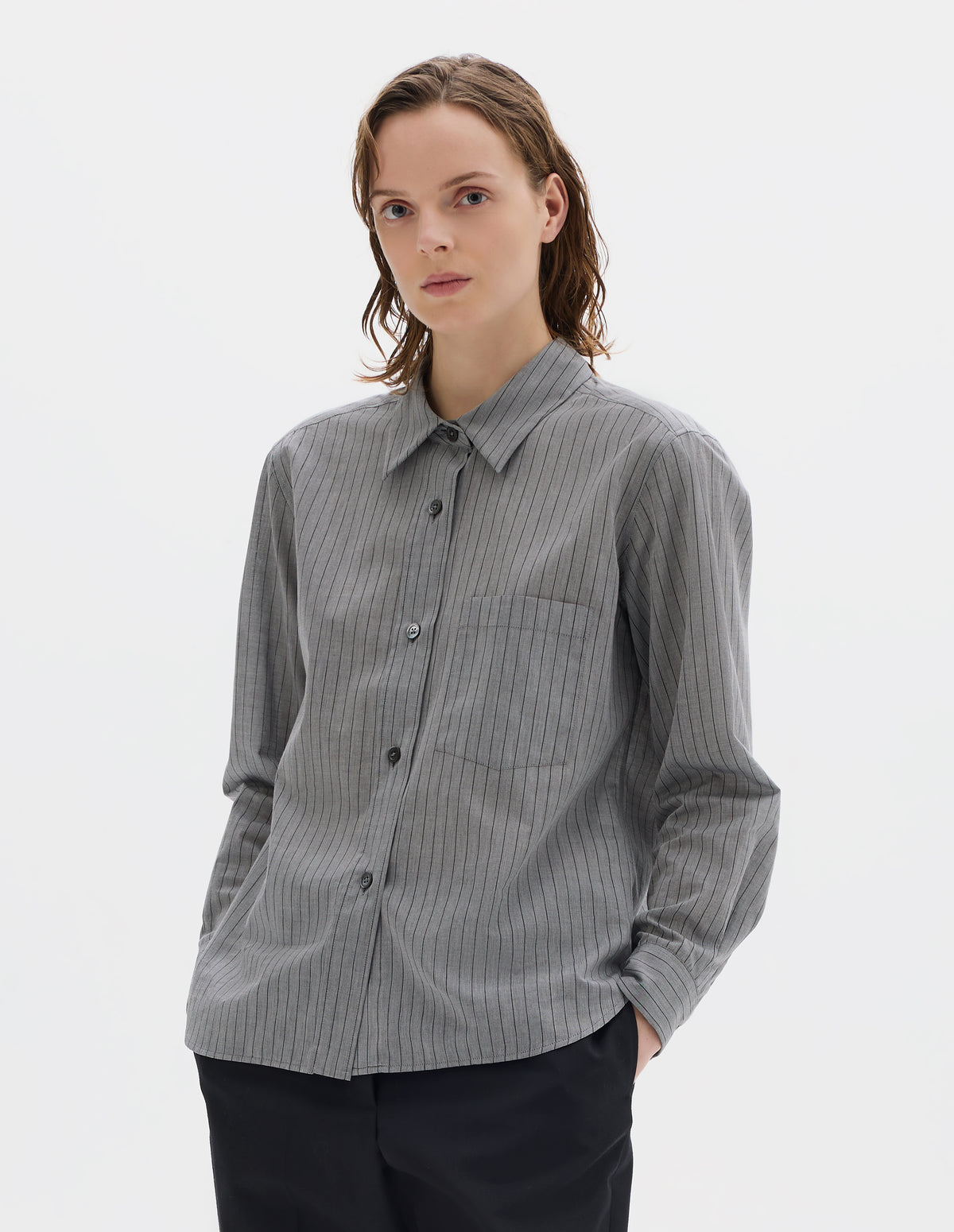 Cropped body image, front view of model wearing grey cotton shirt with black stipes, mother of pearl buttons on raised placket, chest patch pocket and curved hem. Styled with dark blue trousers.