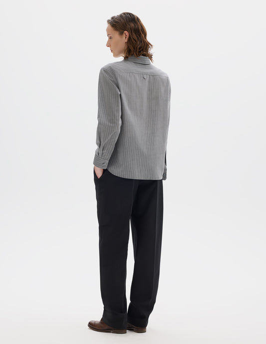 Full body image, back view of model wearing grey cotton shirt with black stipes, hanger loop detail on back yoke, mother of pearl buttons on cuffs and curved hem. Styled with dark blue trousers and brown leather brogues.