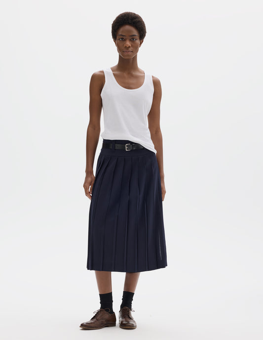 Full body image, front view of model wearing white sleeveless cotton vest with scoop neck. Styled with dark blue skirt with black leather belt, black socks and dark brown leather brogues.