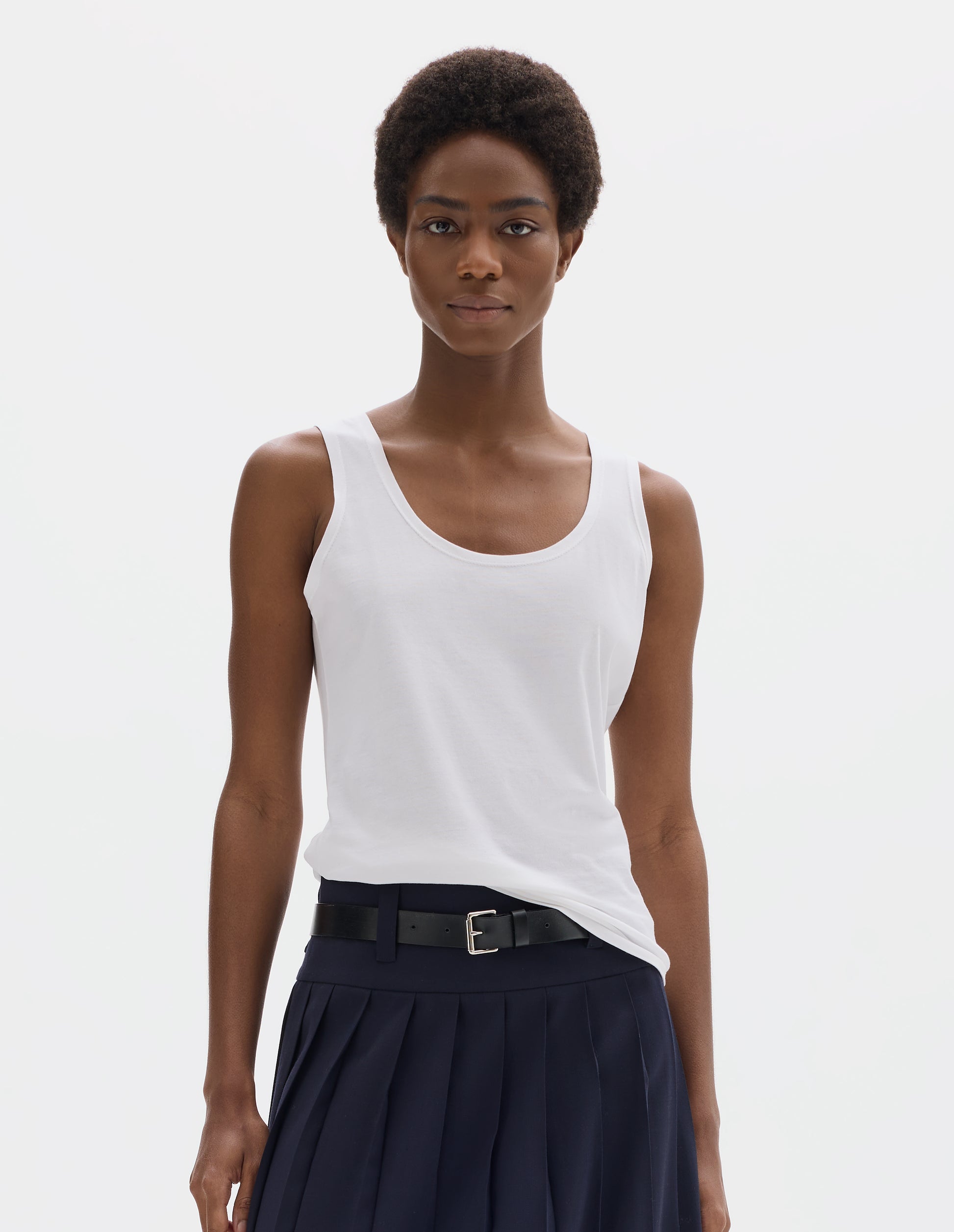 Cropped body image, front view of model wearing white sleeveless cotton vest with scoop neck. Styled with dark blue skirt with black leather belt.