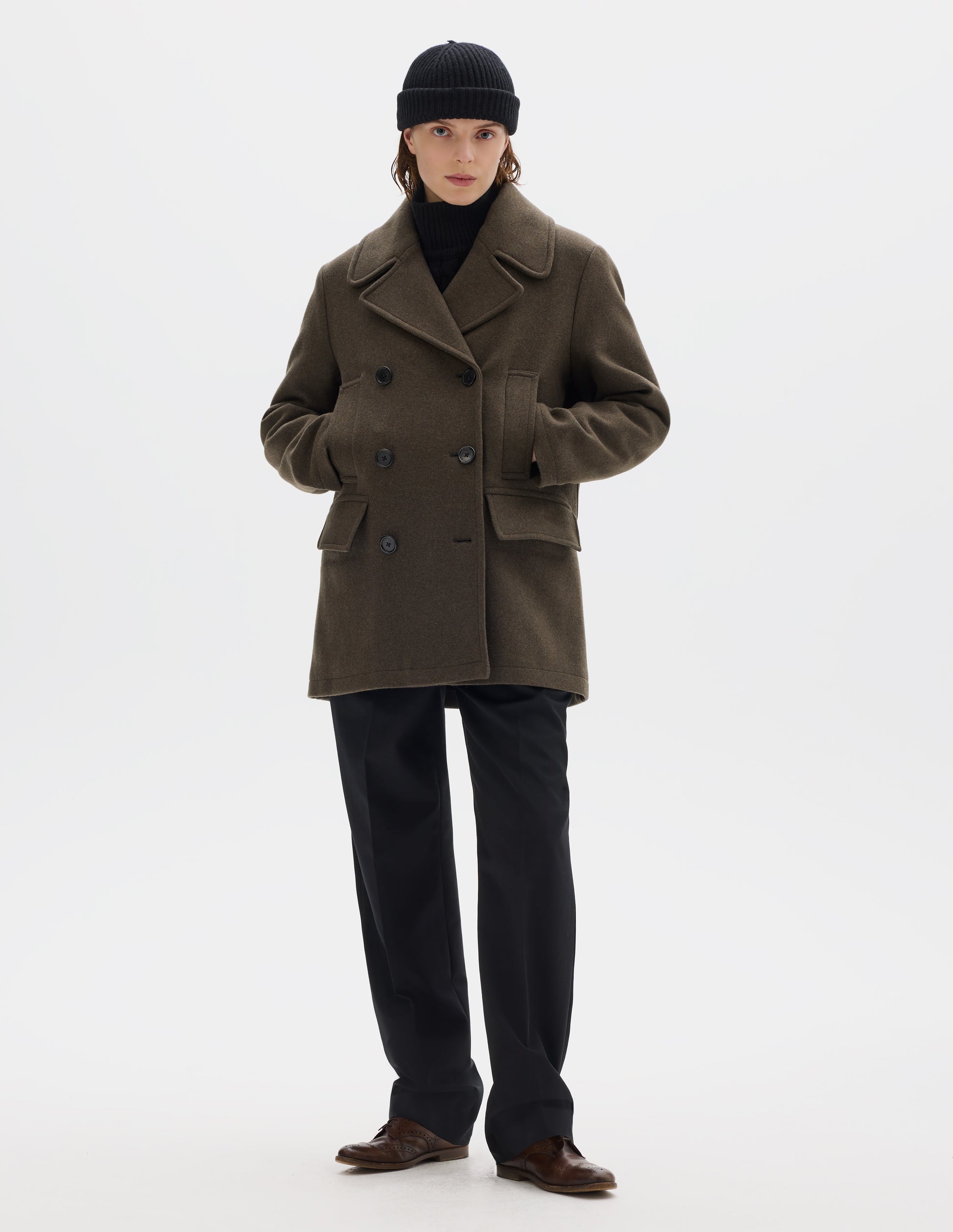 Full body image, front view of model wearing khaki green wool blend coat with large collar and revere, double breasted front with horn buttons, high-set single welt pockets and large flap pockets. Styled with black beanie, black trousers and brown leather brogues.
