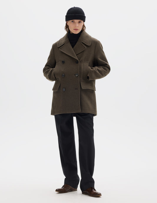 Full body image, front view of model wearing khaki green wool blend coat with large collar and revere, double breasted front with horn buttons, high-set single welt pockets and large flap pockets. Styled with black beanie, black trousers and brown leather brogues.